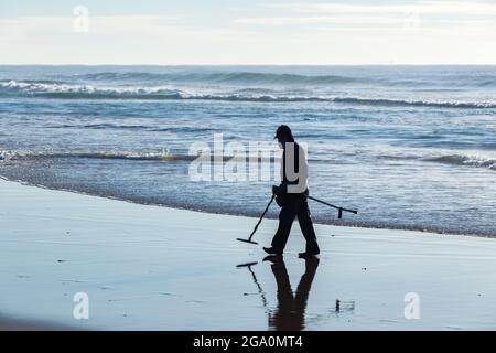 Chasseur de trésors homme non reconnaissable silhoueté marchant sur la plage vagues de l'océan ligne de l'eau avec détecteur de métal à la recherche de trésors perdus. Banque D'Images