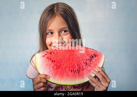 Bon enfant mangeant du melon. Une petite fille mord un gros morceau de pastèque sur fond bleu. Banque D'Images