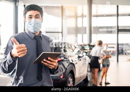 Un saleman asiatique porte un masque chirurgical qui travaille dans un inspecteur vérifiant l'écriture Sur la planchette à pince dans le garage de la concession, attendre le mécanicien dans la salle d'exposition voiture et assurance Banque D'Images