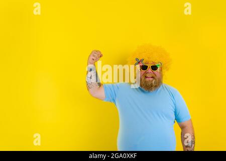 Gros homme avec perruque dans la tête et lunettes de soleil montre son muscle Banque D'Images