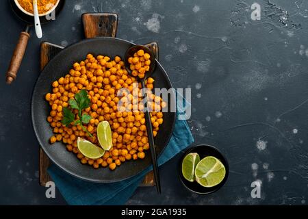 Pois chiches frits au curcuma avec persil et citron vert dans une assiette noire sur un fond de table noir ancien. Pois chiches épicés grillés ou chana ou chole indien, Banque D'Images