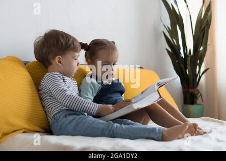 Deux enfants avant-garçonnets lisent un grand livre intéressant de contes de fées sur un lit jaune. Frères et sœurs petits jumeaux lecteur ont plaisir, heureux enfant sur Banque D'Images