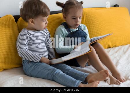 Deux enfants avant-garçonnets lisent un grand livre intéressant de contes de fées sur un lit jaune. Frères et sœurs petits jumeaux lecteur ont plaisir, heureux enfant sur Banque D'Images