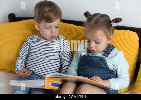 Deux enfants avant-garçonnets lisent un grand livre intéressant de contes de fées sur un lit jaune. Frères et sœurs petits jumeaux lecteur ont plaisir, heureux enfant sur Banque D'Images