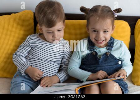 Deux enfants avant-garçonnets lisent un grand livre intéressant de contes de fées sur un lit jaune. Frères et sœurs petits jumeaux lecteur ont plaisir, heureux enfant sur Banque D'Images