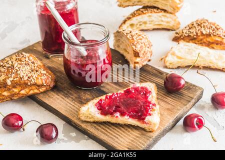 Confiture de cerises maison, cerises fraîches et petits pains de lye coupés sur une planche en bois sur une table blanche Banque D'Images