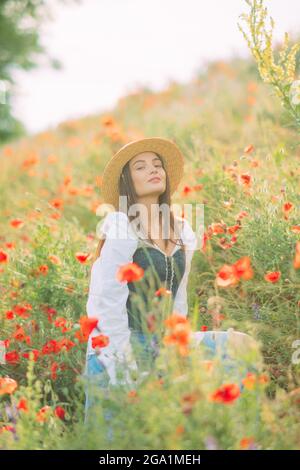 La jeune femme en chapeau de paille apprécie par la floraison des coquelicots parmi les prairies. Banque D'Images