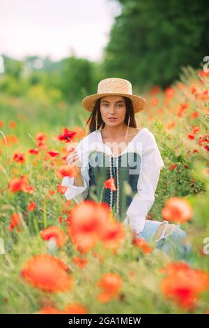 La jeune femme en chapeau de paille apprécie par la floraison des coquelicots parmi les prairies. Banque D'Images