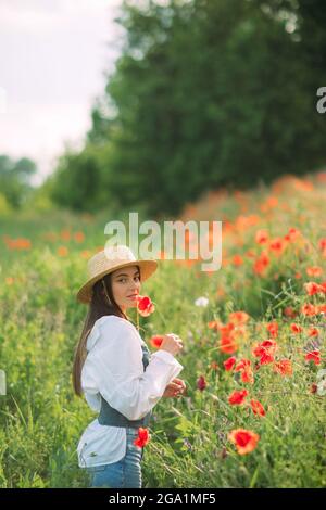 La jeune femme en chapeau de paille apprécie par la floraison des coquelicots parmi les prairies. Banque D'Images