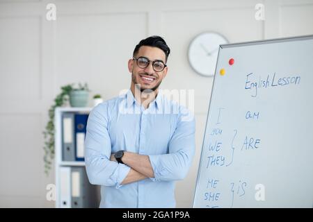 Formation à distance. Joyeux enseignant arabe masculin donnant en ligne classe d'anglais, debout avec les bras croisés près du tableau noir Banque D'Images