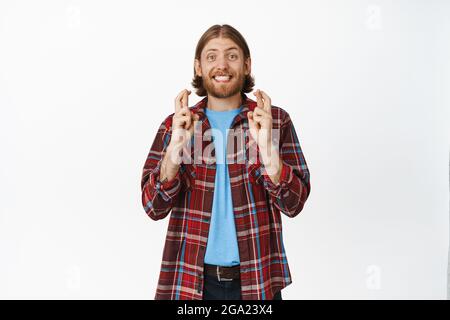 De grands espoirs. Un homme blond excité souriant et regardant ses vœux pieux, croiser les doigts et faire des souhaits, anticipant les grandes nouvelles, l'espoir de gagner, se tenant contre Banque D'Images