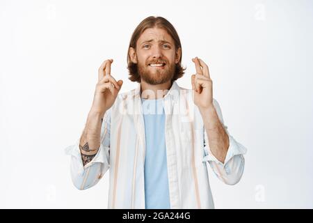 Homme porteur nerveux plein d'espoir dans une chemise, croiser les doigts bonne chance, souhaitant, faire le souhait, anticiper les bonnes nouvelles, attendre les nouvelles, se tenir sur du blanc Banque D'Images