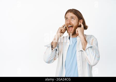 Joyeux blond, qui écoute de la musique avec un casque, des écouteurs sans fil, qui sourit et regarde, chante le long de la chanson, se tient contre le blanc Banque D'Images