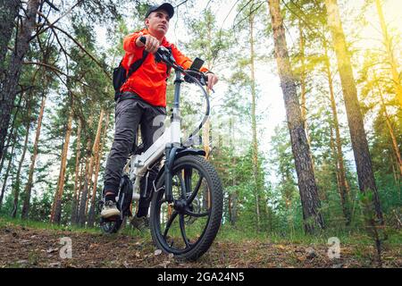 Un homme à vélo électrique dans un parc. Un cycliste se promoit parmi les grands arbres en été au coucher du soleil ou à l'aube. Banque D'Images