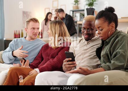 Deux jeunes couples interculturels en vêtements décontractés élégants assis à la rangée sur le canapé et discuter des photos sur les réseaux sociaux en mobile gadgets Banque D'Images