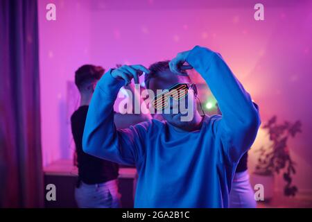 Jeune homme sérieux en vêtements décontractés et lunettes Disco regardant vous vous tenez debout devant ses amis dansant ensemble à la fête à la maison Banque D'Images