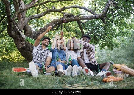 Quatre jeunes dans des vêtements décontractés assis ensemble au jardin vert et en pique-nique d'été. Heureux amis multiculturels boire de la bière froide et manger des saucisses grillées. Banque D'Images