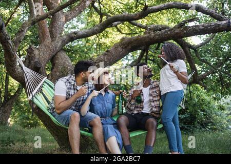 Des amis heureux et variés mangeant des saucisses grillées à la brochette et buvant de la bière tout en étant assis dans un hamac à l'extérieur. Concept de personnes, pique-nique d'été et détente. Banque D'Images