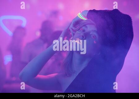 Jeune femme glamour avec des cheveux longs foncés en robe noire vous regardez tout en vous tenant contre l'appareil photo ses amis qui apprécient la fête Banque D'Images