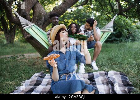 Une jeune femme en tenue d'été boit de la bière et mange des collations tout en étant assise sur un plat dans le jardin. Trois amis multiraciaux qui discutent en arrière-plan dans un hamac. Les jeunes pique-niques. Banque D'Images