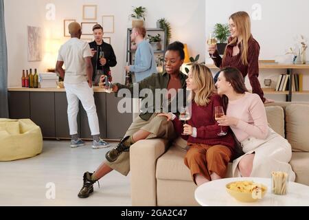 Jeunes femmes interculturelles heureuses avec flûtes de champagne à regarder vous avec des sourires tout en appréciant la fête à la maison contre leurs petits amis Banque D'Images