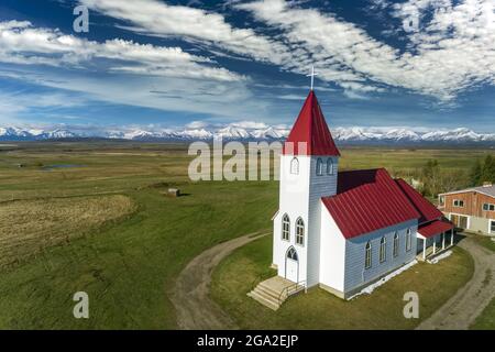 Vue aérienne de l'église au toit rouge, dans une prairie avec des nuages spectaculaires dans un ciel bleu et une chaîne de montagnes enneigée en arrière-plan, nous... Banque D'Images