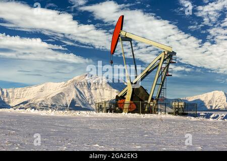 Pumpjack dans un champ enneigé avec des nuages spectaculaires dans un ciel bleu avec une chaîne de montagnes enneigée en arrière-plan; Longview, Alberta, Canada Banque D'Images