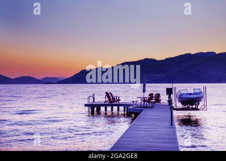 Des chaises Adirondack et une table de pique-nique sont assises sur un quai en bois avec un bateau et un ascenseur sur le lac Okanagan au coucher du soleil; Colombie-Britannique, Canada Banque D'Images