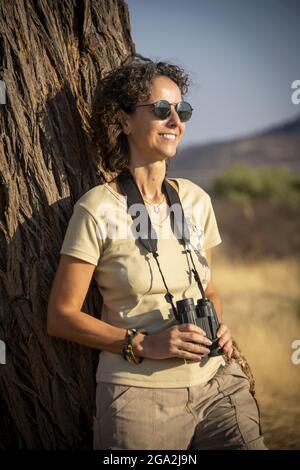 Portrait d'une femme qui s'appuie sur un tronc d'arbre portant un short et des lunettes de soleil et tenant une paire de jumelles tout en regardant dans la lumière du soleil... Banque D'Images