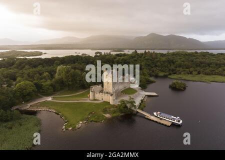 Vue aérienne du château de Ross sur une crique de Lough Leane avec un ciel gris brumeux et des montagnes silhouetées en arrière-plan au parc national de Killarney Banque D'Images