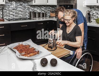 Une femme paraplégique qui prépare un repas pour sa famille dans sa cuisine tout en travaillant à partir d'un fauteuil roulant quand son mari l'encadre; Edmonton, Alberta, Canada Banque D'Images