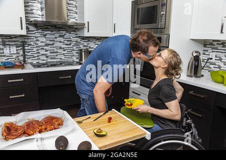 Une femme paraplégique préparant un repas pour sa famille dans sa cuisine tout en travaillant à partir d'un fauteuil roulant quand son mari l'embrasse Banque D'Images