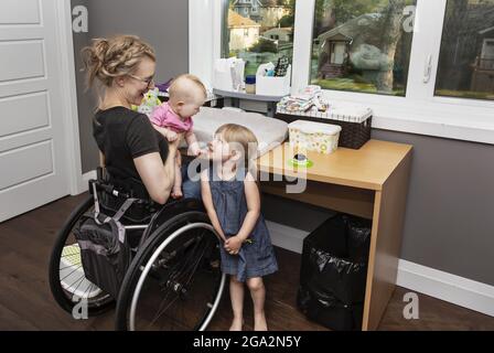 Une mère en fauteuil roulant change la couche de son bébé, tandis que la grande sœur aide à l'amuser; Edmonton, Alberta, Canada Banque D'Images