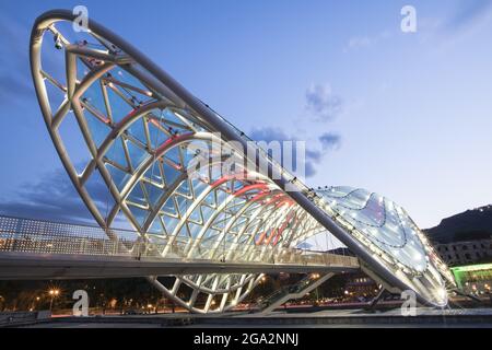 Le pont piétonnier en forme d'arc de la paix, illuminé au crépuscule, enjambant la rivière Mtkvari (Kura) reliant le parc Rike et le vieux Tbilissi Banque D'Images