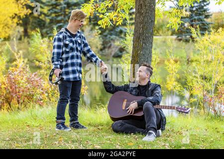 Un jeune homme avec le syndrome de Down fait une bosse de poing après avoir joué un tambourin tandis que son père a joué une guitare dans un parc de ville lors d'une chaude soirée d'automne Banque D'Images