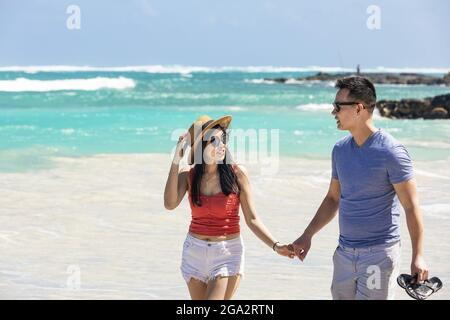 Un couple marchant sur une plage de sable blanc tenant les mains et parlant ensemble tout en appréciant des vacances tropicales le long de la côte d'Oahu, Hawaï Banque D'Images