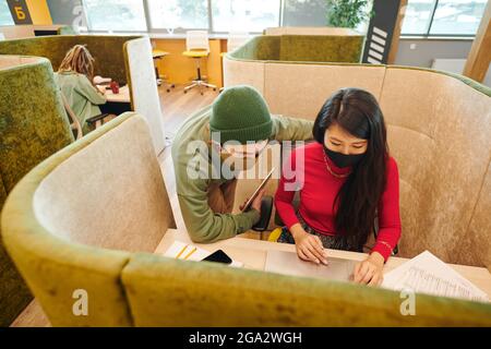 Jeune femme d'affaires asiatique en vêtements décontractés et masque de protection agitant main tout en regardant le couple heureux sur l'écran d'ordinateur portable Banque D'Images