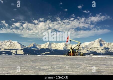 Pumpjack dans un champ enneigé avec des nuages spectaculaires dans un ciel bleu avec une chaîne de montagnes enneigée en arrière-plan; Longview, Alberta, Canada Banque D'Images