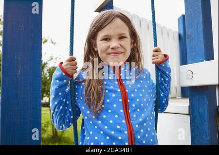 Jeune fille assise sur une balançoire sur un terrain de jeu, souriant à la caméra; Allemagne Banque D'Images