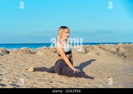 Jeune femme de forme physique en sport porter s'étirant assis sur la ficelle dans la plage de sable pendant l'entraînement en plein air. Mode de vie actif et soins de santé. Copier Banque D'Images