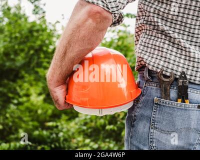 Homme attrayant dans des vêtements de travail, tenant le casque de construction dans ses mains contre le fond des arbres, le ciel bleu et le coucher du soleil. Vue de l'arrière. Laboratoire Banque D'Images