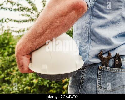 Homme attrayant dans des vêtements de travail, tenant le casque de construction dans ses mains contre le fond des arbres, le ciel bleu et le coucher du soleil. Vue de l'arrière. Laboratoire Banque D'Images