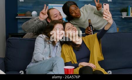 Groupe de personnes de race mixte prenant des photos avec le téléphone tout en étant assis sur un canapé dans le salon passant du temps ensemble. Amis multiraciaux poster selfie sur le partage d'Internet avec d'autres personnes Banque D'Images