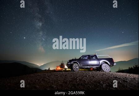 Voiture de sport noire garée sur la route de montagne de terre près du camp touristique avec tente orange sous ciel étoilé de nuit, sur fond de silhouettes de montagne Banque D'Images