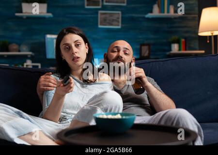 Un jeune couple qui passe la soirée ensemble vêtu de pyjamas en train de passer du temps ensemble, une femme qui change des programmes télévisés en utilisant la télécommande pour se détendre sur un canapé en mangeant du pop-corn, en buvant de la bière Banque D'Images