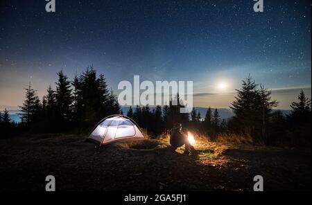 Vue latérale d'un jeune homme assis sur l'herbe près de sa tente éclairée et regardant les étoiles de nuit et la lune briller. Loisirs dans les montagnes camping en plein air. Banque D'Images