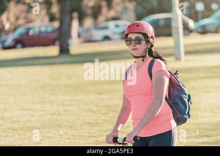 Portrait d'une jeune femme à bord d'un scooter de poussée à travers les parcs d'Adélaïde tout en portant des lunettes de soleil avec un haut rose et un casque de vélo Banque D'Images