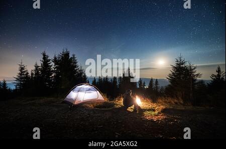 Vue latérale d'un jeune homme assis sur l'herbe près de sa tente éclairée et regardant les étoiles de nuit et la lune briller. Loisirs dans les montagnes camping en plein air. Beauté du ciel nocturne de l'étoile lunaire. Banque D'Images