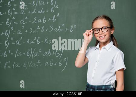 joyeux élève ajustant des lunettes près du tableau noir avec des équations mathématiques Banque D'Images