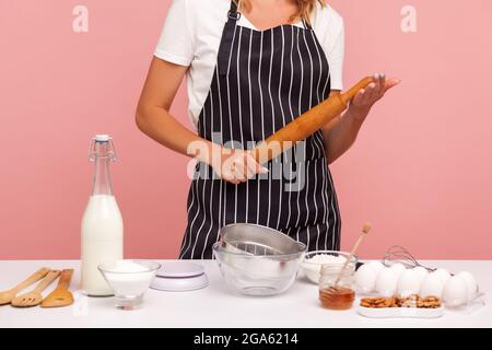 Image rognée de boulanger dans un tablier rayé, tenant la goupille dans les mains, étant entouré d'ingrédients pour faire des pâtisseries, cuire des gâteaux. Goujon intérieur Banque D'Images